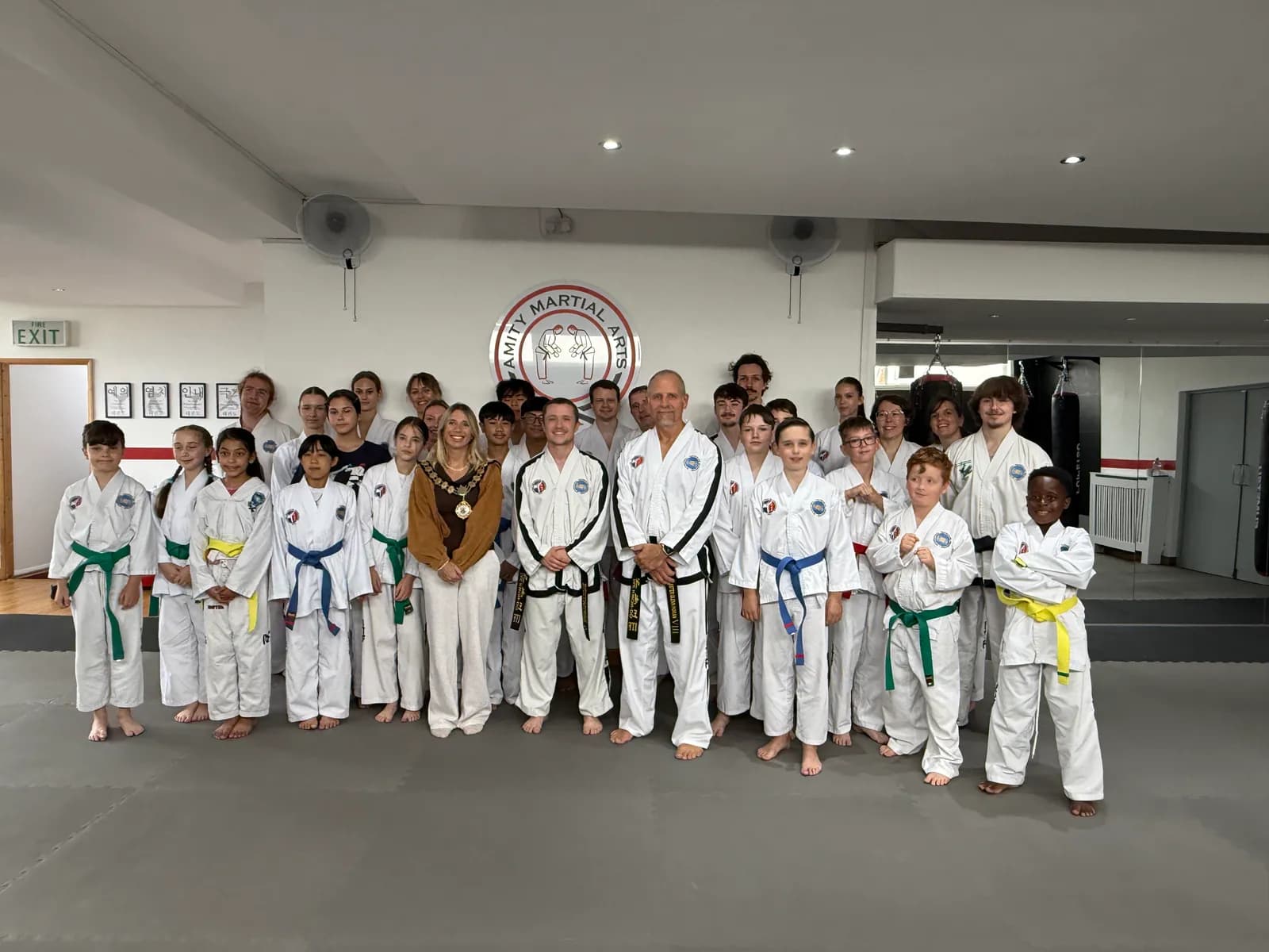 A group of diverse children and adults wearing taekwondo uniforms, some with colored belts, pose for a group photo in a martial arts studio with a woman in a brown and white outfit.