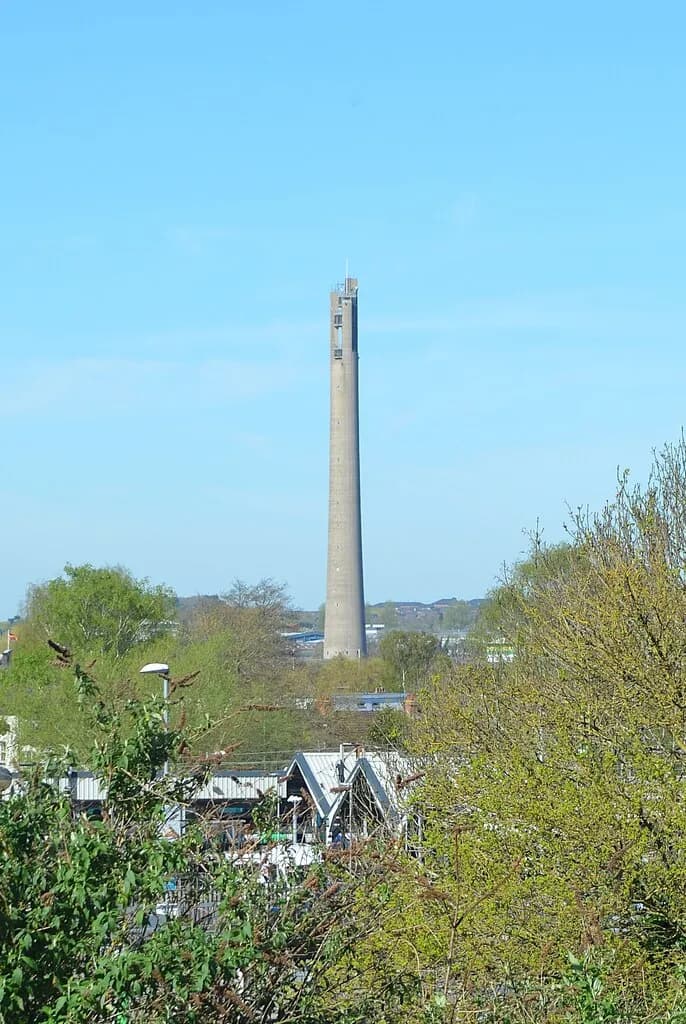 A tall, narrow concrete tower with small windows near the top stands prominently against a clear blue sky, surrounded by green trees and distant buildings.