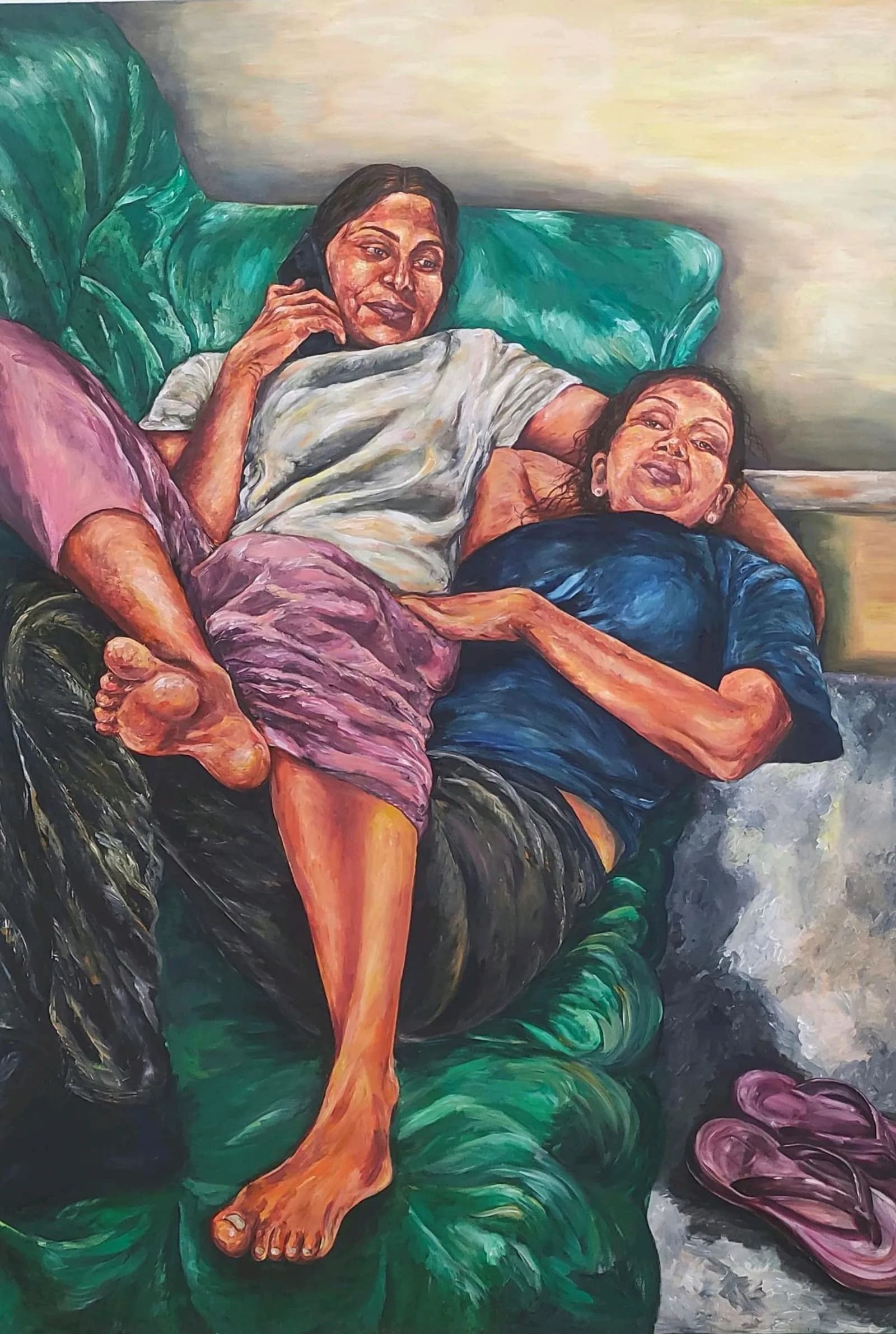 Two women reclining on a green sofa, one talking on a phone and the other resting her head on the first woman's arm.