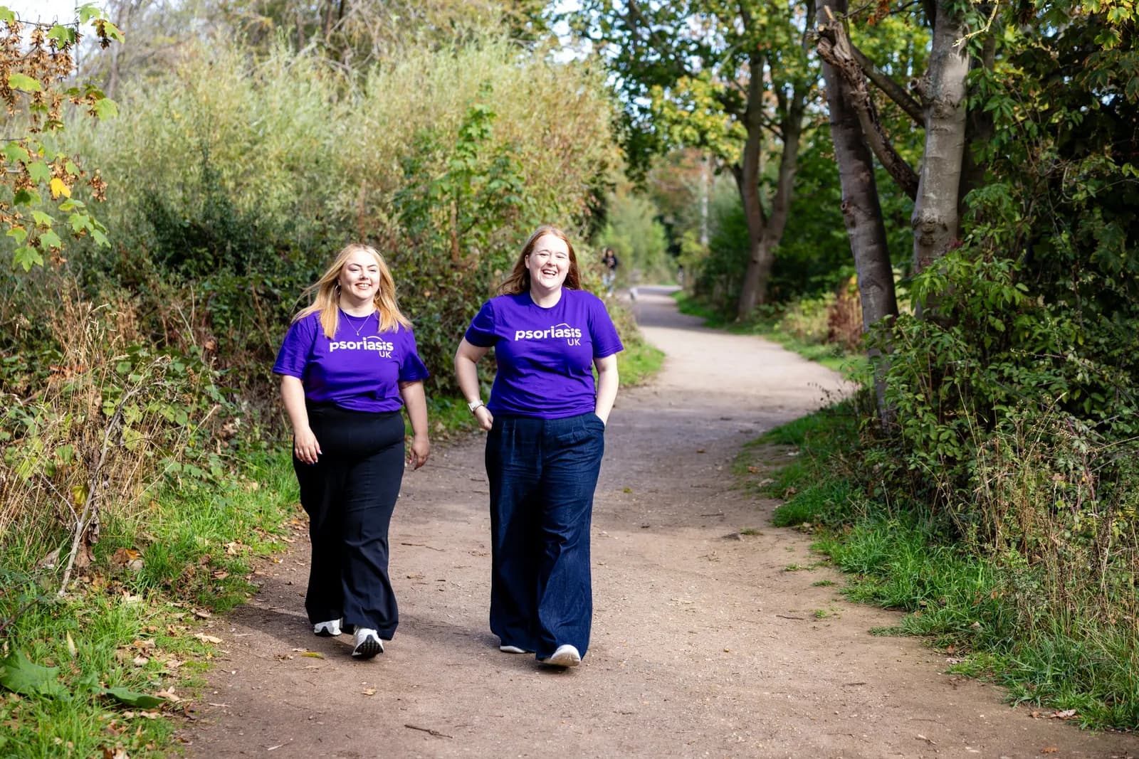 Two women wearing purple t-shirts with 'Psoriasis UK' written on them are smiling and walking on a dirt path surrounded by trees and bushes.